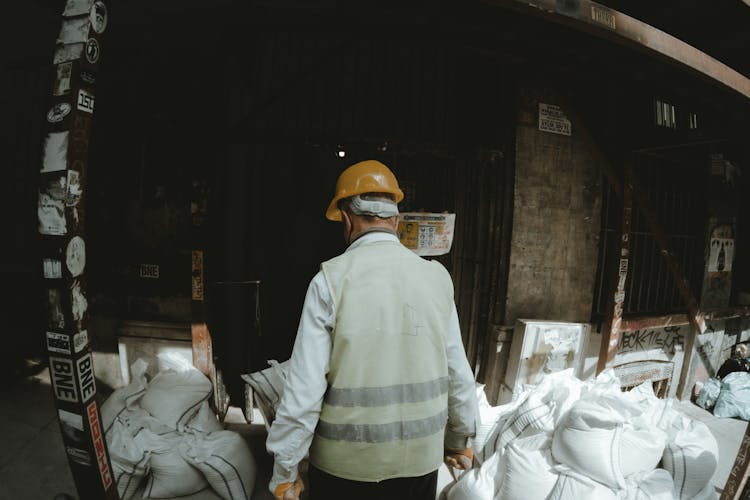 Back View Shot Of A Person Wearing Reflective Vest And Hard Hat Standing Near White Sacks