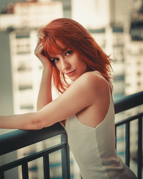 Free Woman in a white tank top posing on a balcony with a blurred cityscape background Stock Photo
