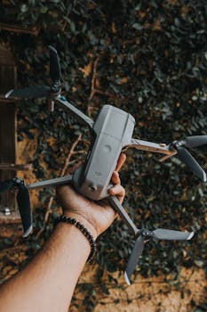 Close-up of a hand holding a DJI drone, set against a leafy backdrop outdoors.