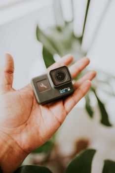 A hand holding a compact action camera with green leaves in the background.