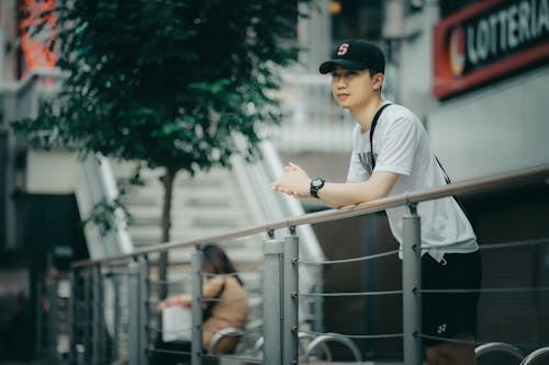 A Man in Gray Shirt and Black Cap Leaning by the Metal Railings while Looking Afar