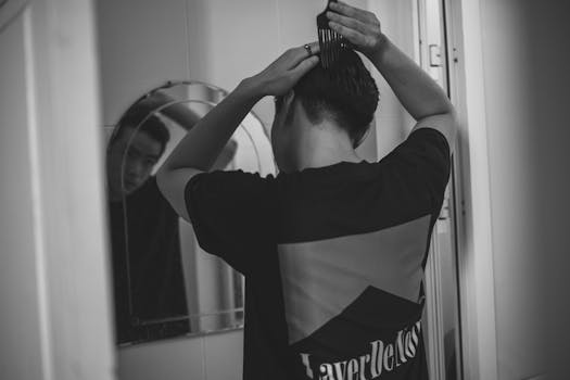 Monochrome image of a man grooming, combing hair in front of a mirror indoors.