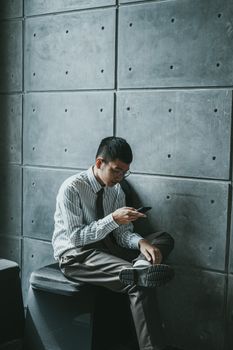 A young professional texting while seated against an industrial concrete wall.