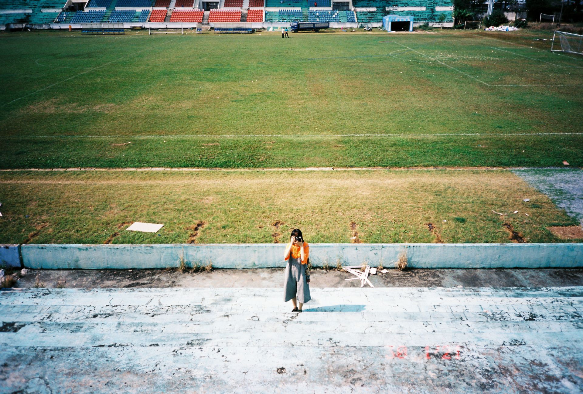 A lone woman takes a photo on empty bleachers with a vast sports field in the background.