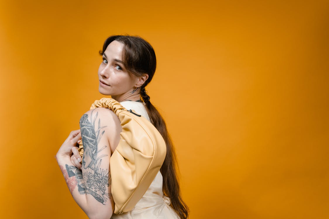 Tattooed Woman Carrying Cute Handbag Near Orange Wall while Looking at the Camera