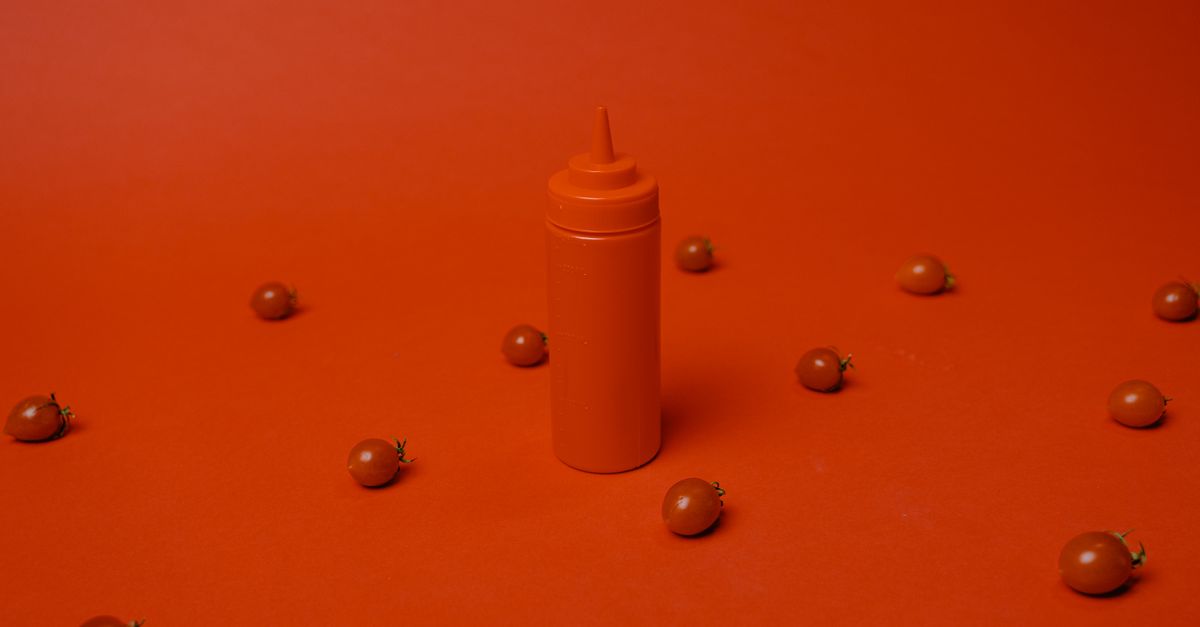 Garrafa vermelha de ketchup cercada por tomates cereja em fundo vermelho. Simples e ousado.