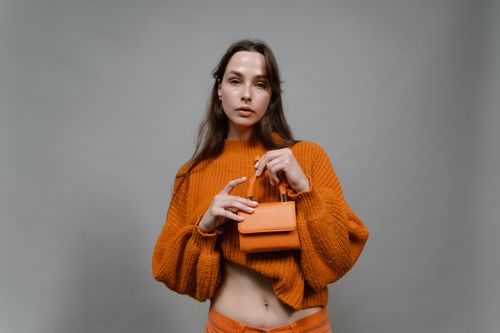 Free Portrait of a stylish woman wearing an orange sweater and holding a matching handbag against a gray background. Stock Photo