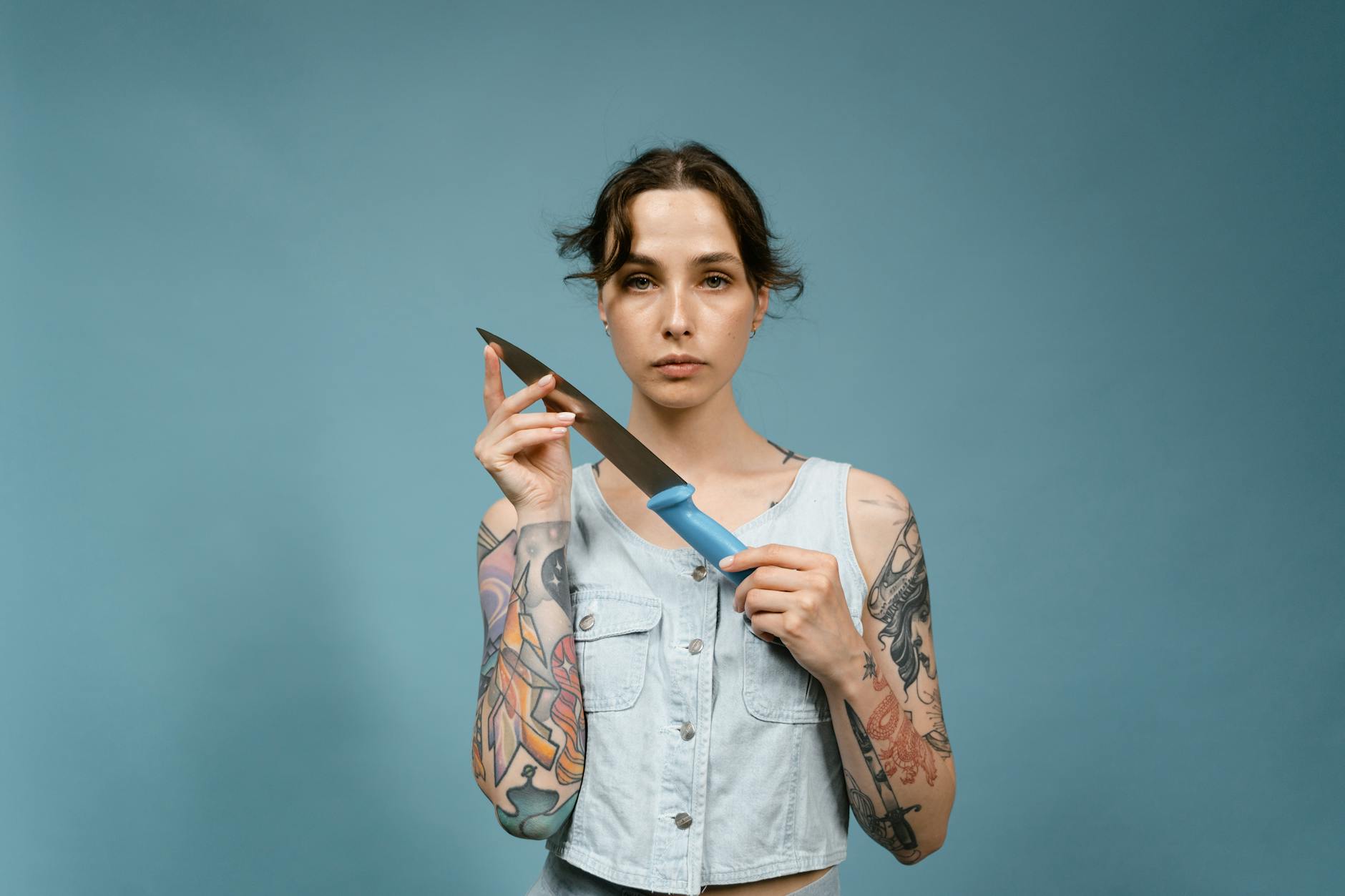 Portrait of a tattooed woman holding a knife against a blue background in a studio setting.