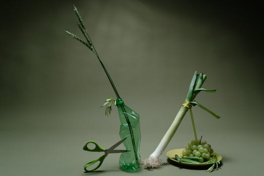A creative still life featuring leeks, grapes, and a plastic bottle on a green background.