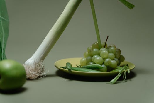 A minimalist arrangement of green grapes, leeks, and an apple on a plate.