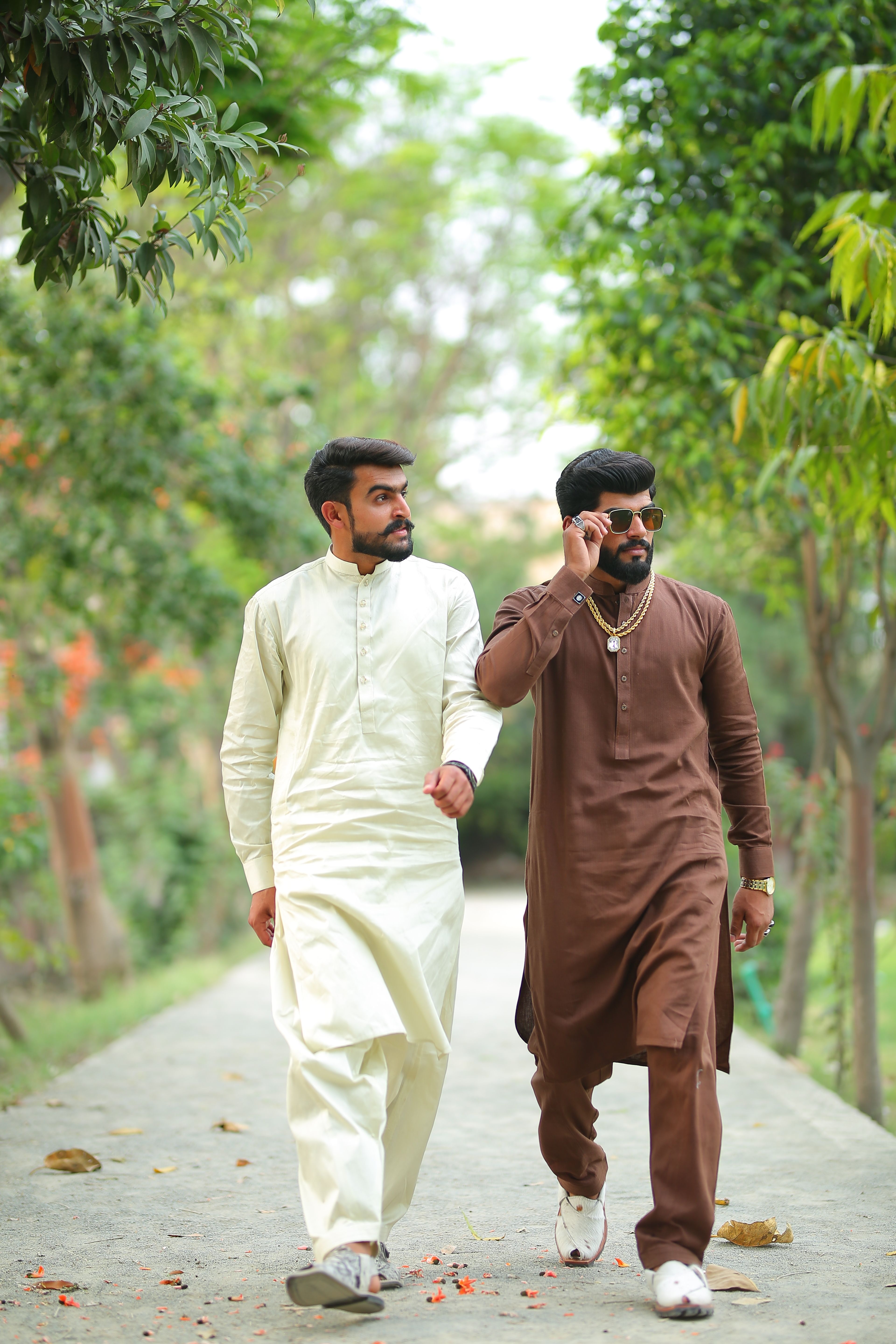 Two stylish men in traditional shalwar kameez walk confidently in a lush park setting.