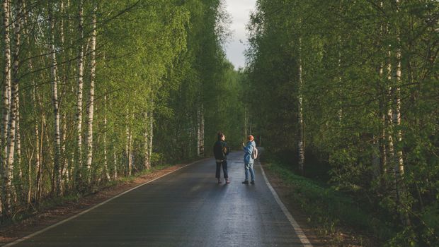 Two individuals strolling along a tree-lined road, surrounded by lush green forest.