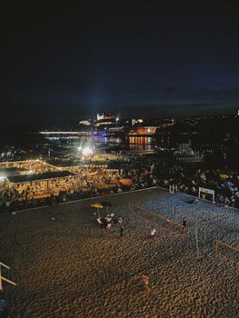 Crowded beach at night with lively festival atmosphere and volleyball games.