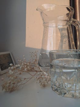 A serene still life featuring a glass pitcher, water glass, dried flowers, and digital clock.