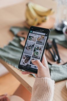 Close-up of a woman's hand holding a smartphone, browsing culinary images online, indoors.