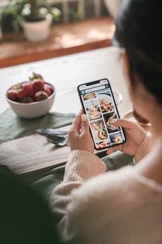 Woman using smartphone to browse food recipes in a cozy kitchen setting with strawberries nearby.