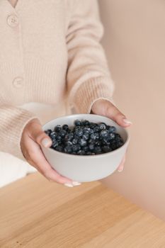 A woman in a cozy sweater holds a ceramic bowl full of fresh blueberries indoors.
