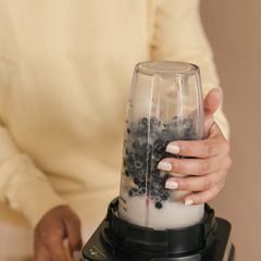Hands holding a tall blender cup of blueberries and milk on a black motor base, against a cream background