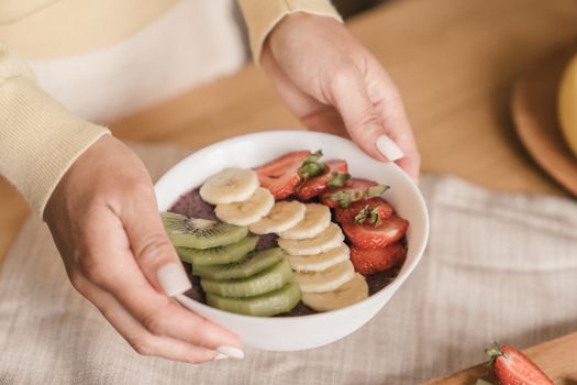 Delicious smoothie bowl topped with fresh fruits like kiwi, banana, and strawberries, held by manicured hands.
