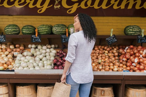 Free Woman in White Long Sleeve Shirt and Blue Denim Jeans Standing in Front of Fruit Stand Stock Photo
