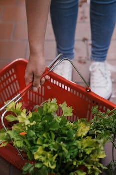 A hand putting fresh green vegetables in a red shopping basket indoors.