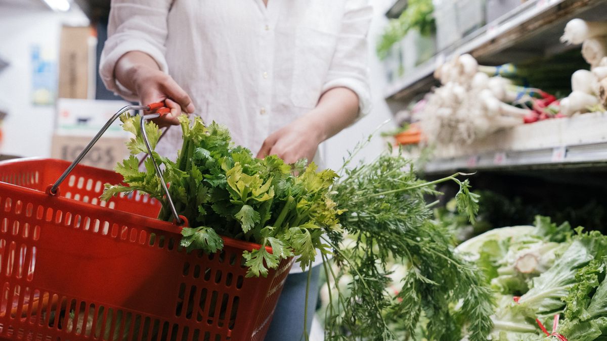 A person holding a grocery basket filled with fresh greens like celery and parsley in a store.