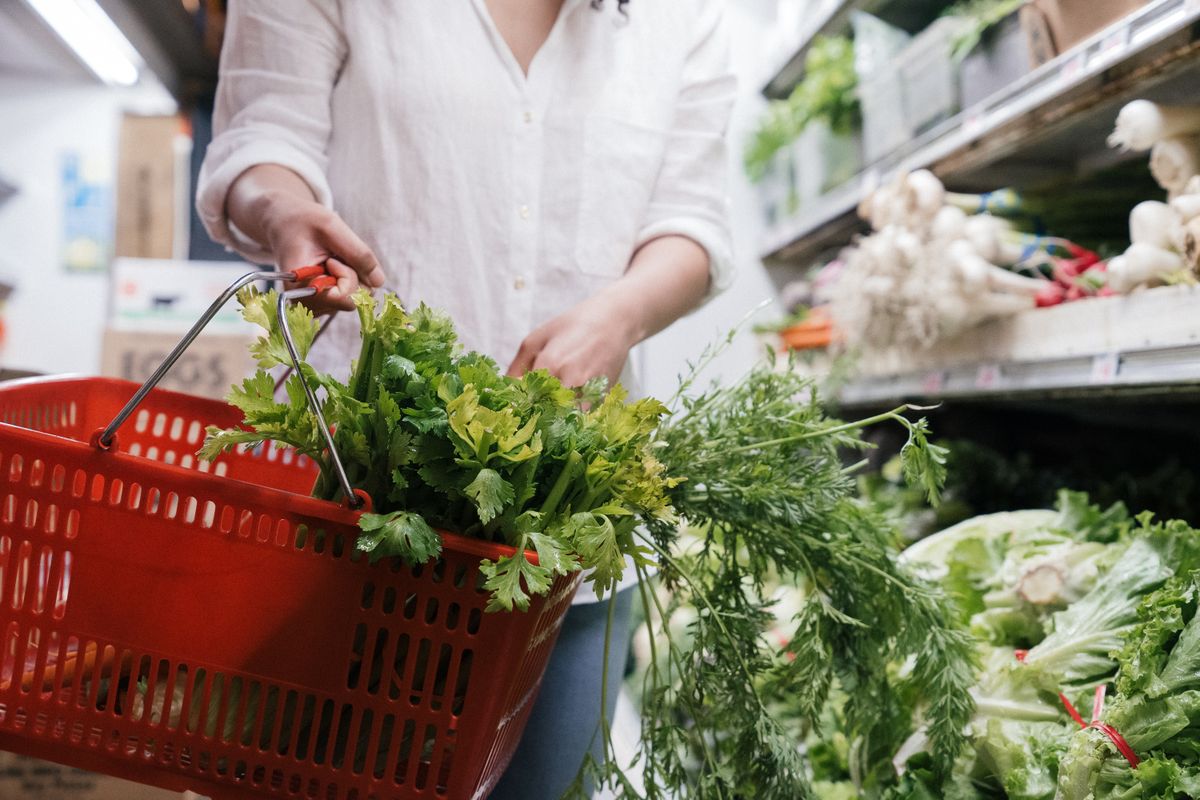 A person holding a grocery basket filled with fresh greens like celery and parsley in a store.