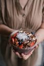 A close-up image of a hands holding an acai bowl topped with fresh fruits, coconut, and chocolate drizzle.