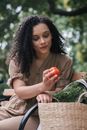 A Woman in Brown Blouse Looking at the Tomato she is Holding while Sitting on a Wooden Bench