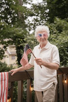 Senior man in patriotic sunglasses holding American flag outdoors on Fourth of July.