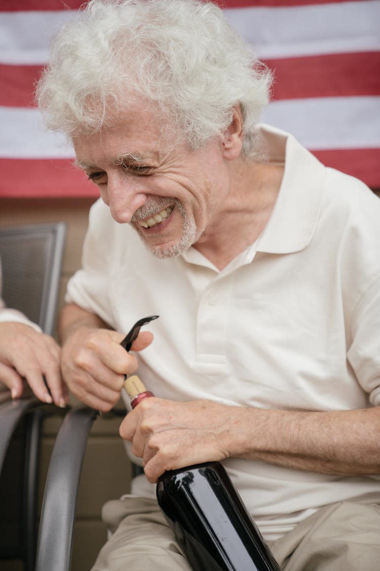 Cheerful Senior Man Sitting In Chair And Opening Wine Bottle