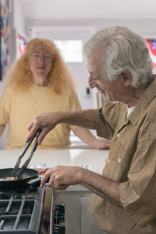 Free Elderly man cooking on stove while woman watches in a bright home kitchen setting. Stock Photo