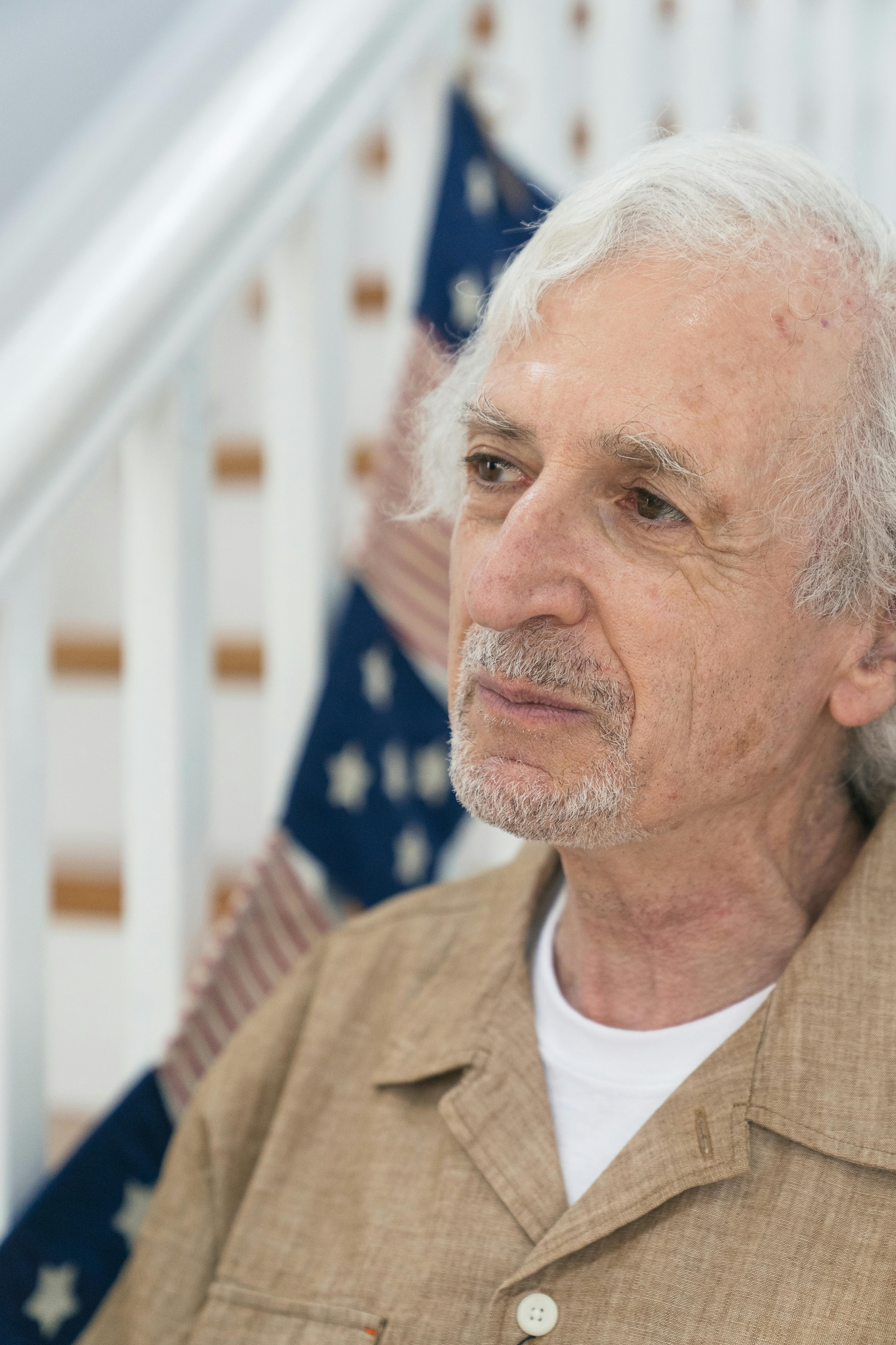 Close-up portrait of a senior man with white hair, reflecting beside an American flag backdrop.