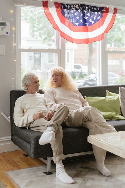 Free Elderly LGBT couple enjoying a peaceful day at home, sitting on a sofa under a US flag. Stock Photo