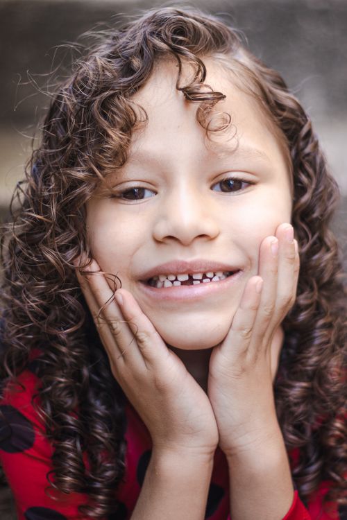 Free Charming close-up of a young girl with curly hair smiling at the camera, hands on face. Stock Photo