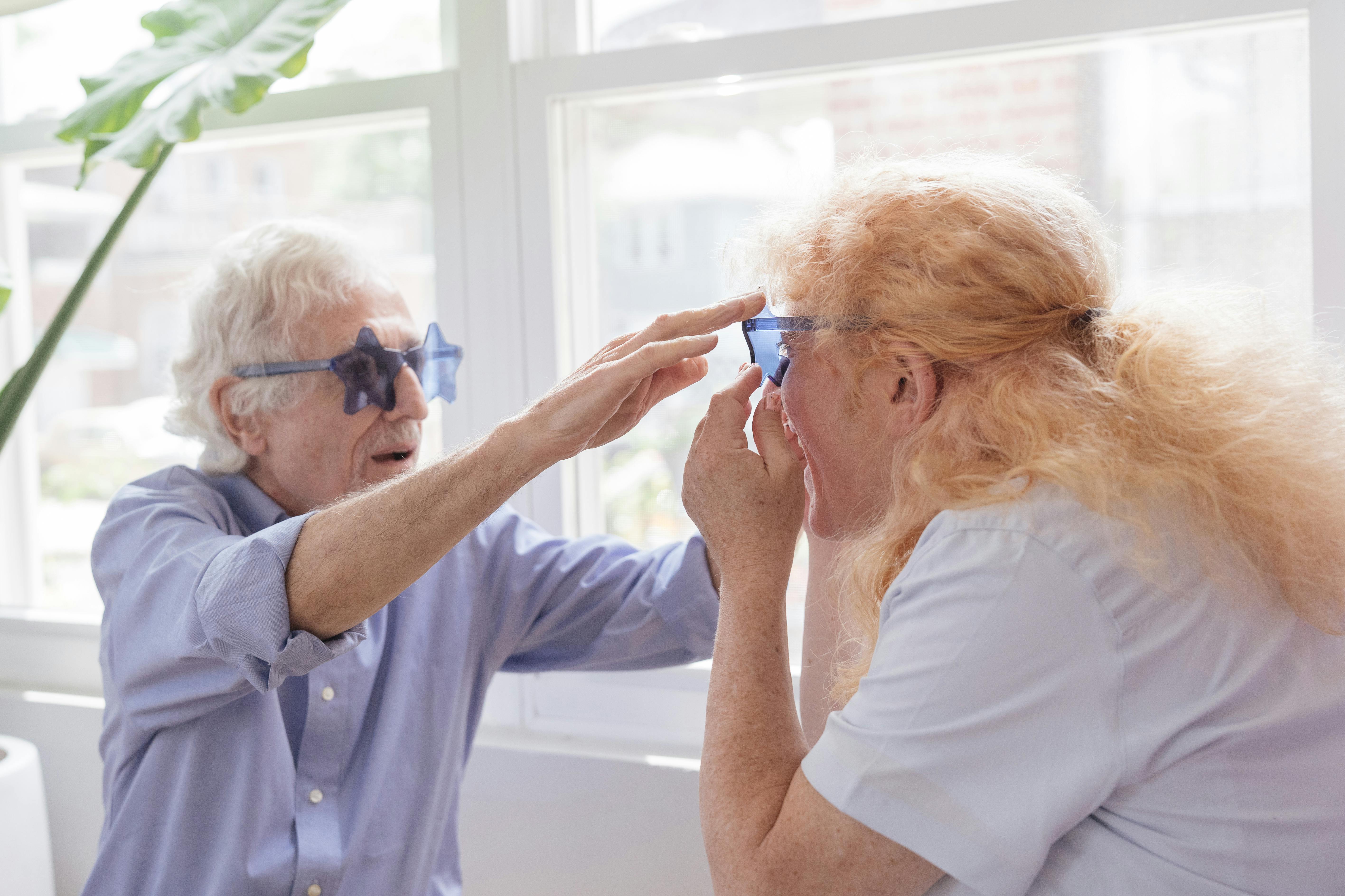 Elderly couple joyfully laughing and wearing fun star-shaped glasses indoors in natural light.