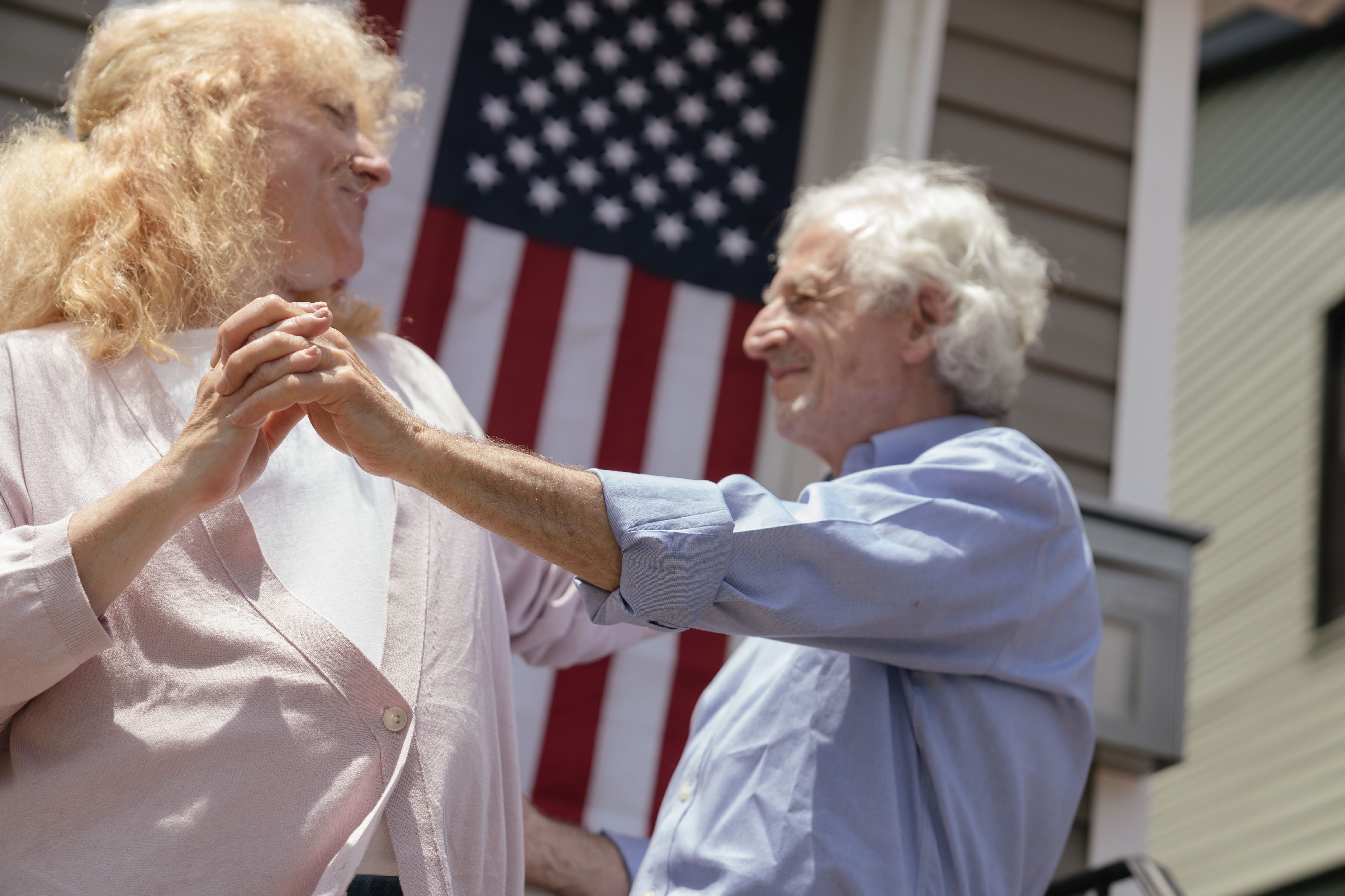 Smiling senior couple dancing with an American flag background, celebrating Independence Day.