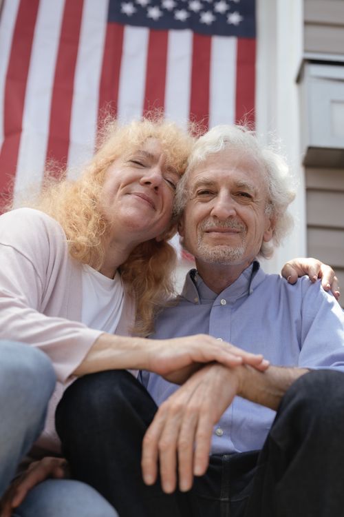 Free Smiling elderly couple seated together in front of American flag, embracing happiness. Stock Photo
