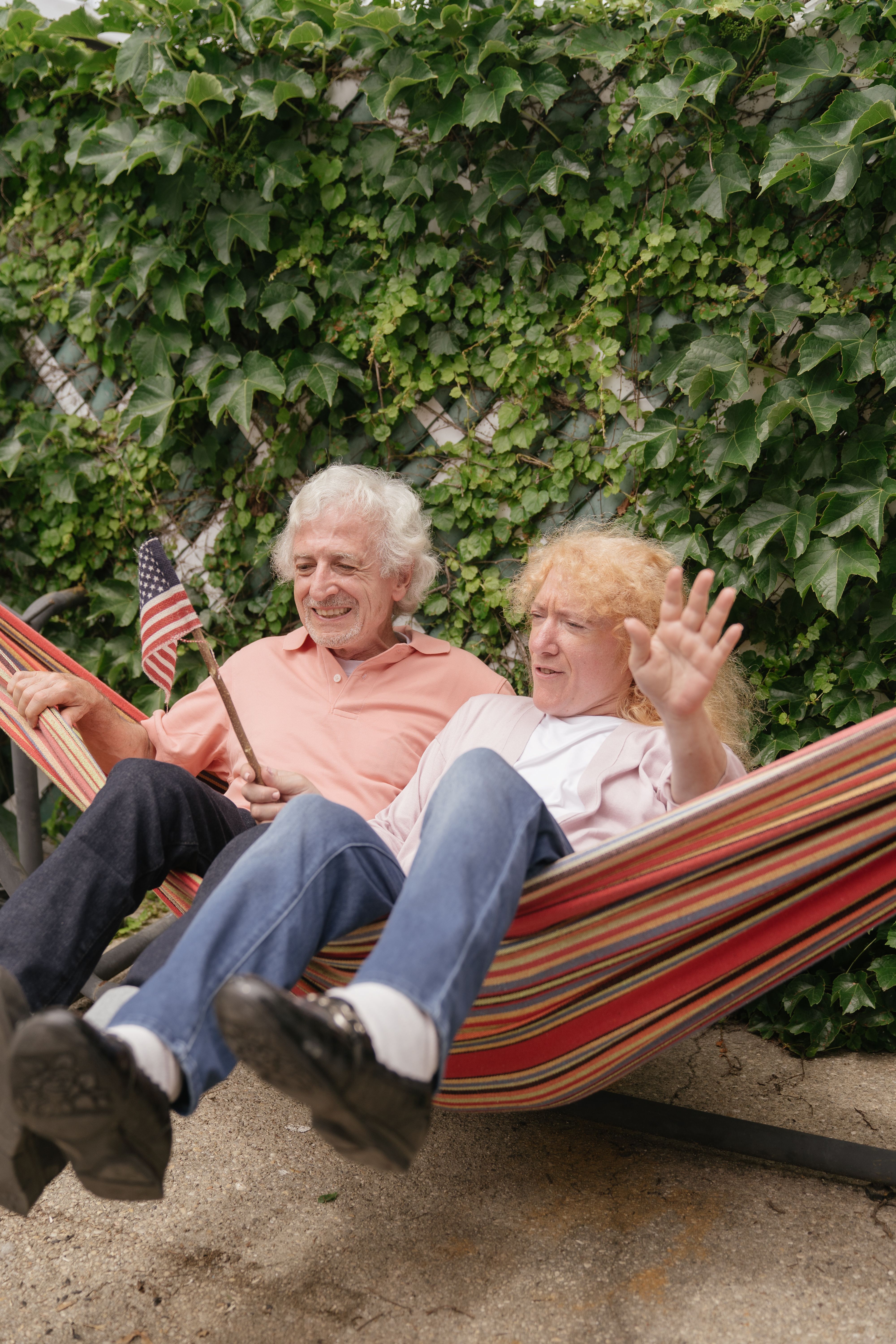 Happy senior couple sitting in a hammock waving, surrounded by lush greenery.