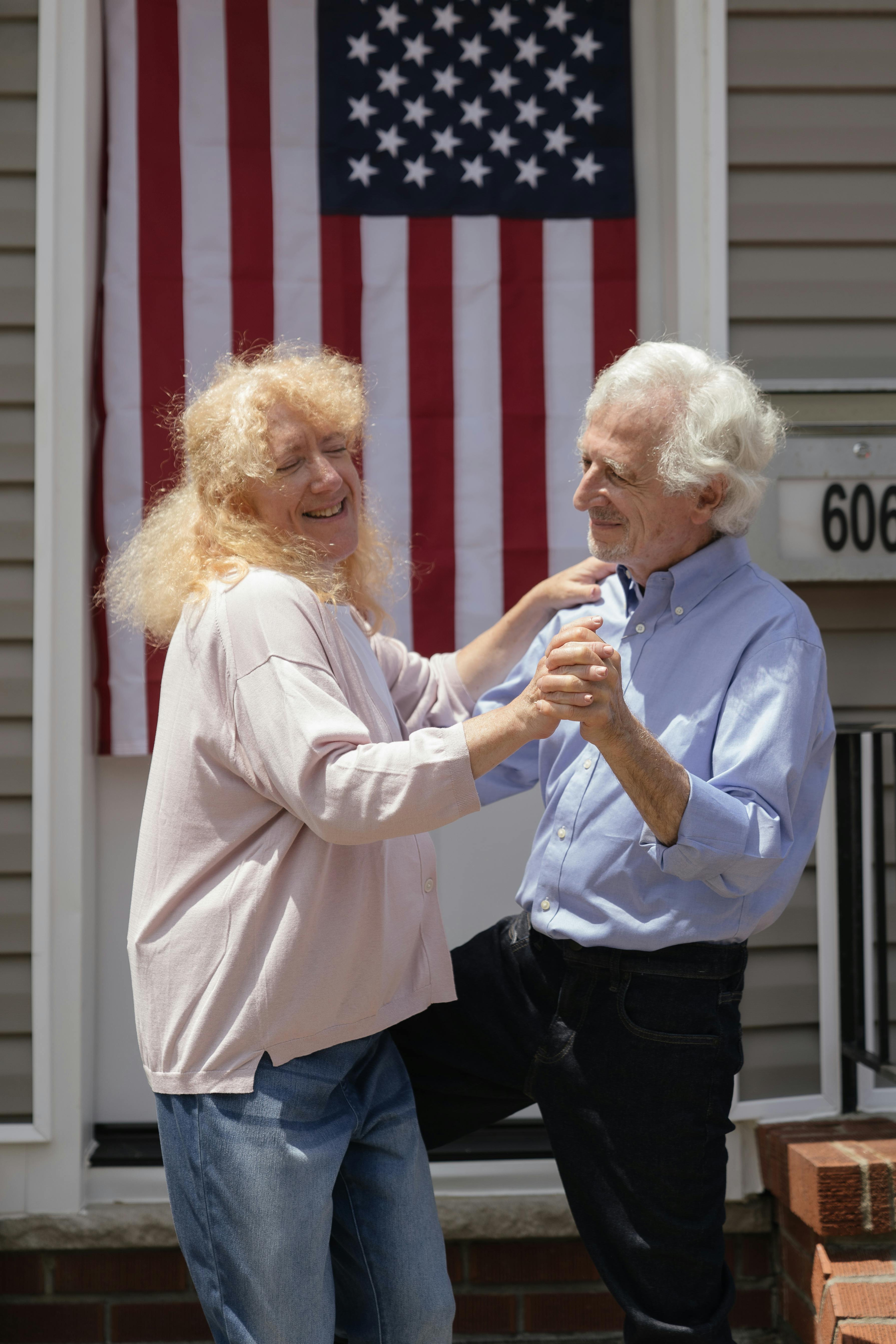 Happy senior couple dancing outside with American flag backdrop on a sunny day.