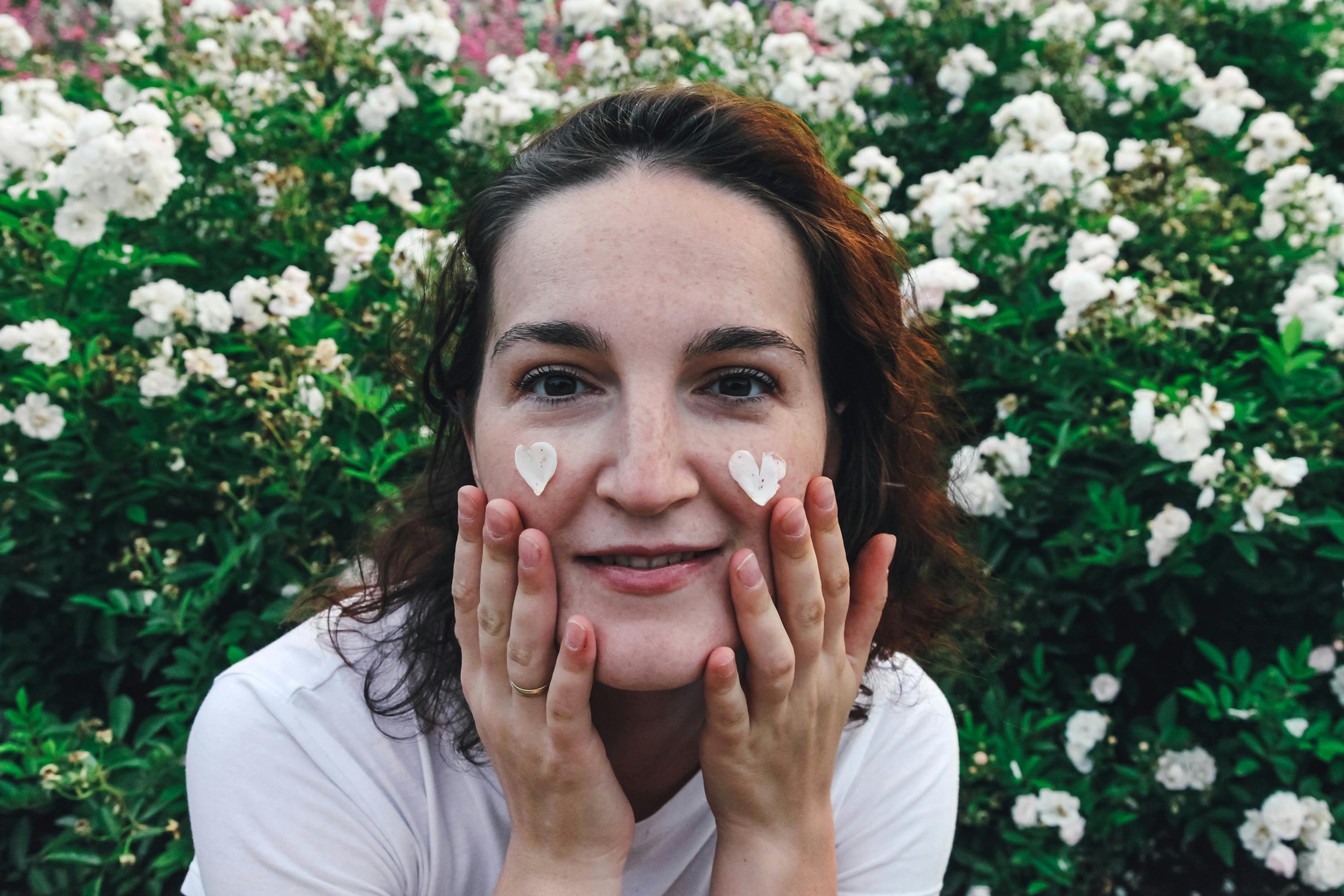 A woman in a blossoming white flower garden applying skincare cream with a heart shape.