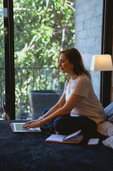 Elderly woman sitting on bed using laptop indoors with natural light.
