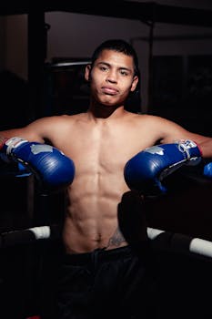 Young male boxer with blue gloves posing confidently in a dimly lit gym setting.