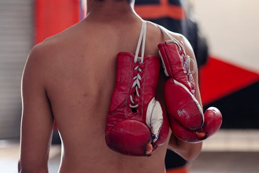 Shirtless boxer with red gloves hanging on shoulder, ready for training.