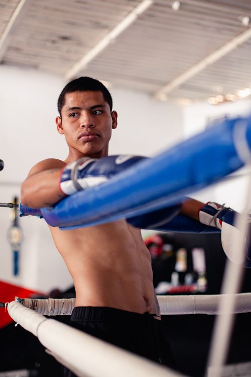 Free Shirtless young boxer resting in the corner of a gym boxing ring, wearing blue boxing gloves. Stock Photo