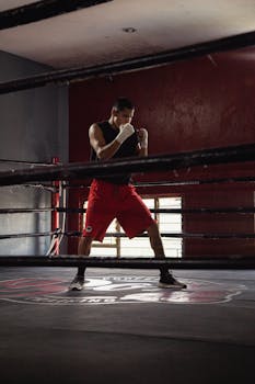Focused male boxer in red shorts practices inside a boxing ring at a gym.