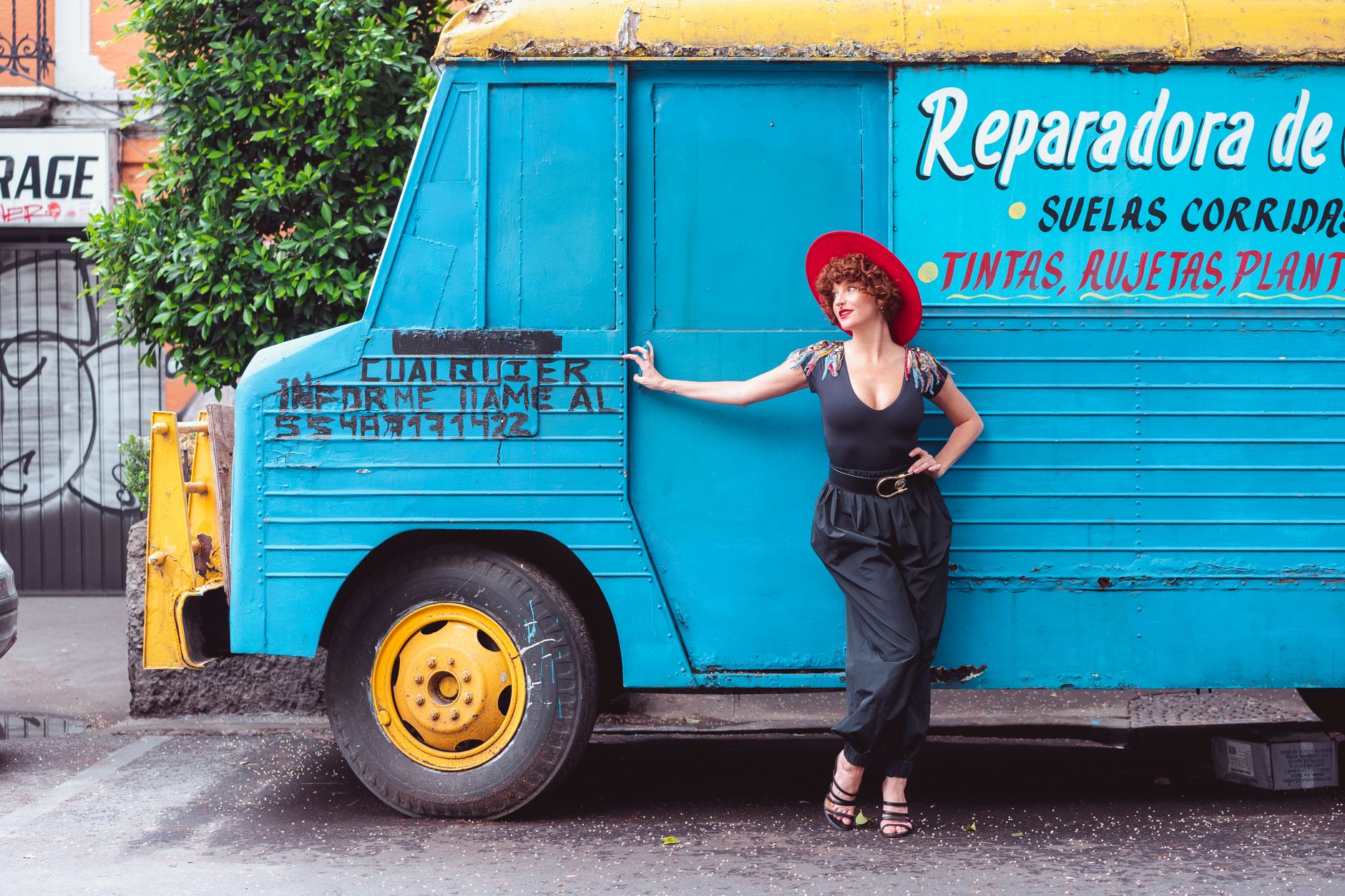 Stylish woman in red hat and black outfit posing by a vintage blue truck with Mexican signage.