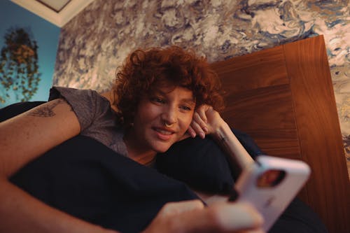 A Woman in Gray Shirt Lying on the Bed while Using Her Mobile Phone