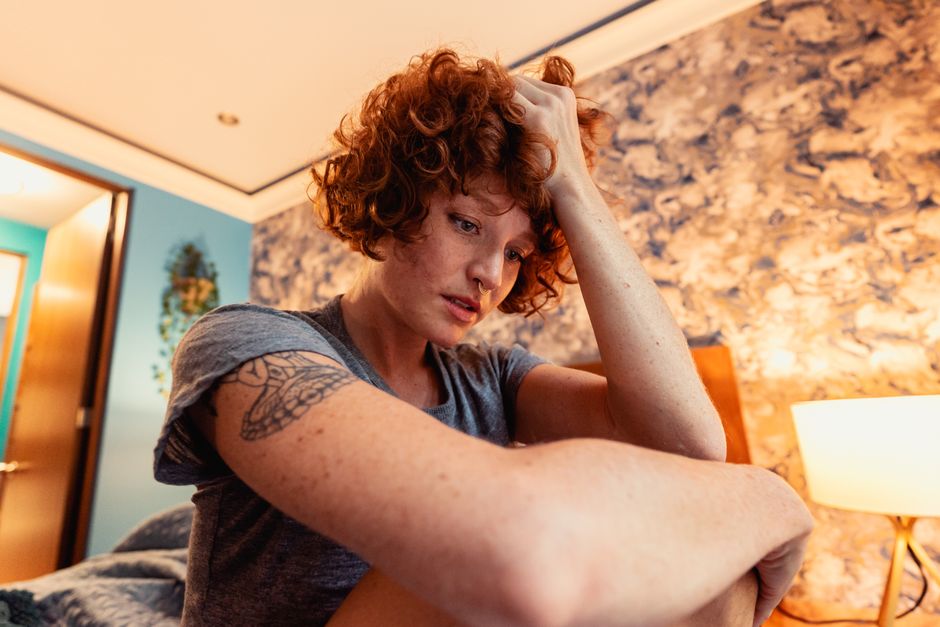 A contemplative woman with curly red hair sits indoors in a warmly lit room.