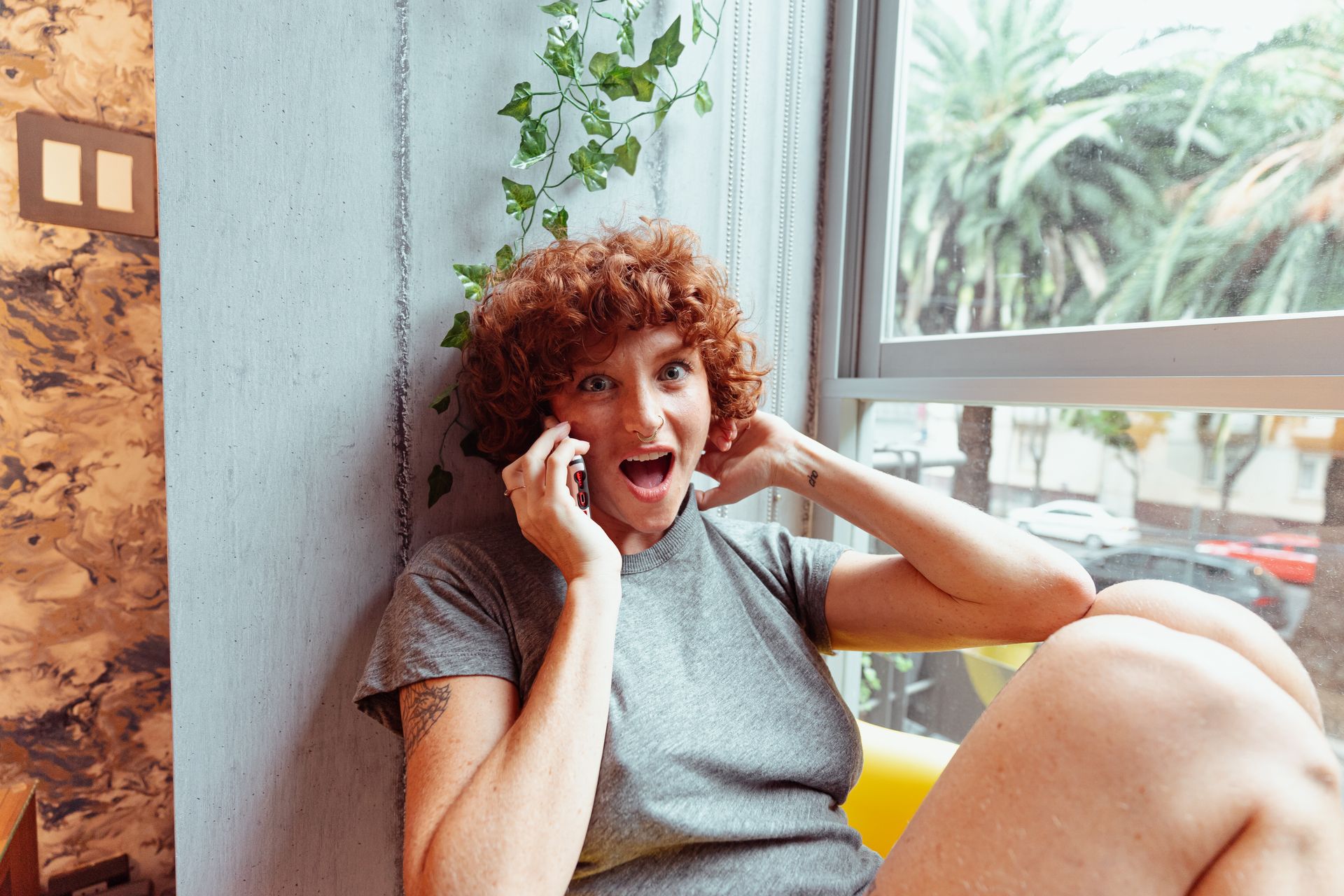 Caucasian woman with curly red hair expressing shock while on the phone indoors by a window.